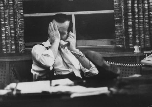 A man sitting at a desk discussing a deal on the phone for a job he doesn't like.