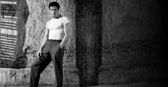 A man in a fitted, tucked-in white t-shirt and dark pants stands casually against a textured wall with one hand in his pocket, in a black-and-white photo.